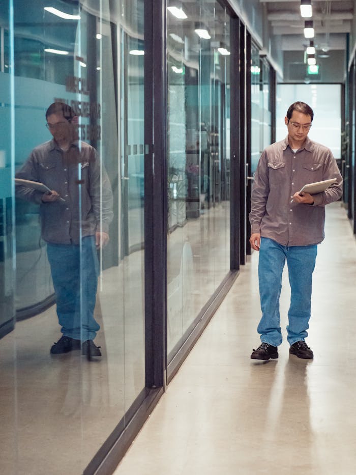 Asian man in casual attire walking through a modern office hallway holding a laptop.