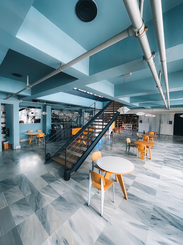 Interior of bright modern cafe with tables and chairs near stairway with railing with pipes on ceiling